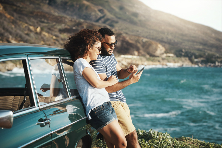 A man and woman leaning on a car checking app on phone.