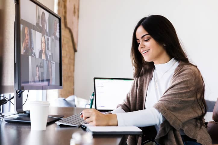 Woman at her desk writing notes while on a video group conference displaying on her computer screen.