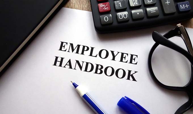 A paper with a heading of employee handbook, surrounding by glasses, pens, notepad, calculator.
