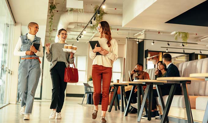 Group of female employees walking in the office holding coffee and notebooks.