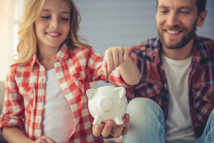 Daddy holding a piggybank while the daughter puts the coin inside.