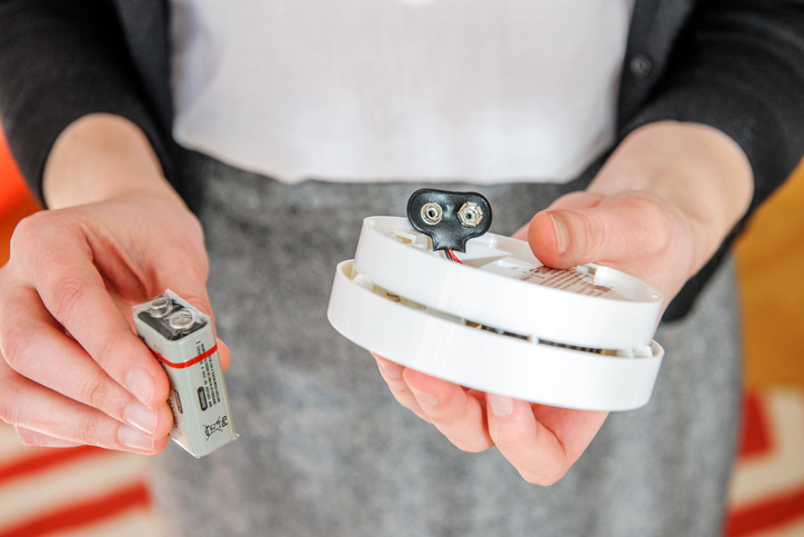 A lady holding a battery and smoke detector in both hands.