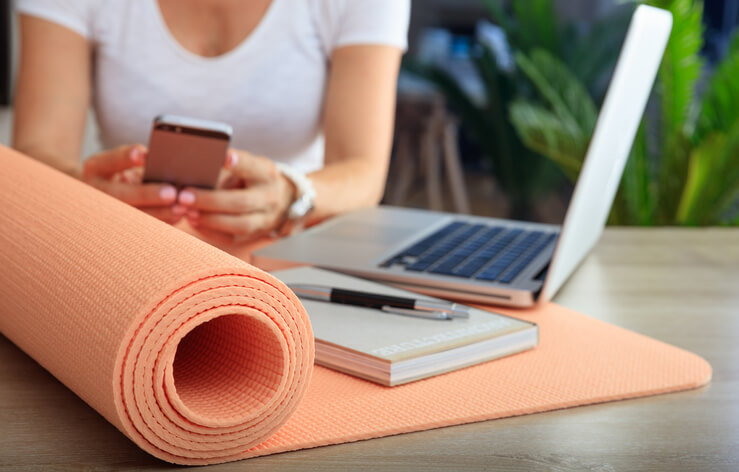 Woman using her smartphone as her laptop and notepad lay on a yoga mat.