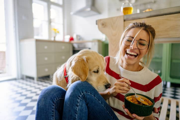 A woman sitting on the floor eating pasta while trying to avoid her dog.