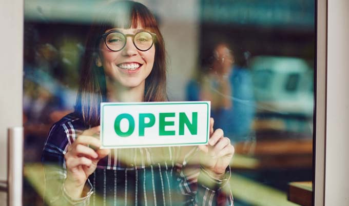Woman holding a OPEN sign in front of glass door.